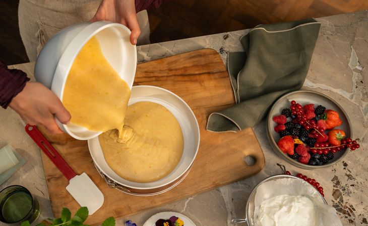 Eine Person gießt hellen Kuchenteig aus einer Rührschüssel in eine runde Backform auf einem Holzbrett, umgeben von frischen Beeren, Schlagsahne, einem Spatel, Kräutern und essbaren Blumen auf einer Arbeitsplatte aus Marmor. 