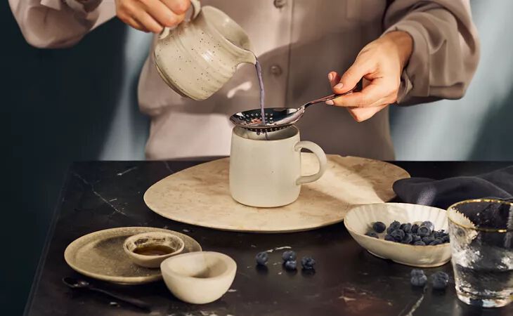 Eine Person, die warme Mondmilch durch ein Sieb in einen Becher gießt, der von Blaubeeren und beruhigenden Zutaten umgeben ist.