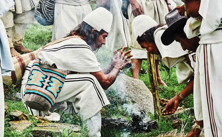 Un groupe de personnes portant des vêtements blancs traditionnels avec des accessoires tissés sont rassemblées autour d’un petit feu en extérieur, sur l’herbe verte. Une personne est agenouillée et met ses mains devant le feu, peut-être dans un geste cérémonial, tandis que les autres l’observent ou participent. 