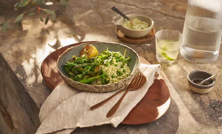 Eine Keramikschale mit einem Orzo-Risotto mit grünem Gemüse – Erbsen, Brokkoli, Spargel und Rucola. Serviert mit einer gegrillten Zitronenhälfte. 