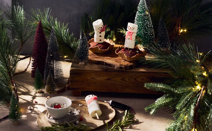 Świąteczne, czerwone ciastka z bałwankami z pianek marshmallow w scenerii przypominającej las.