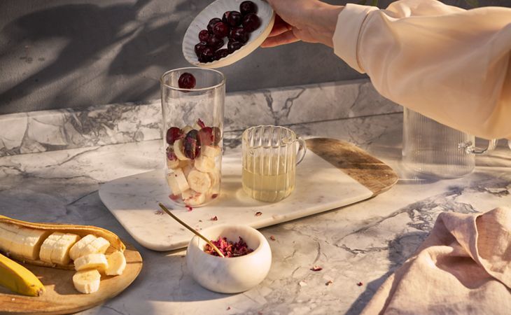Personne ajoutant des cerises dans un verre contenant des tranches de banane et du jus de citron pour préparer un smoothie nourrissant pour la peau.