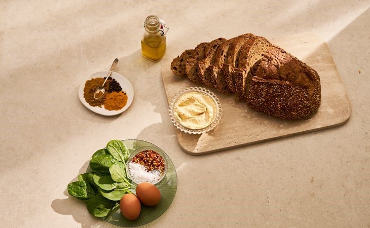 Die Zutaten für einen proteinreichen vegetarischen Toast: Körnerbrot, Hummus, Spinat, Gewürze, Eier und Olivenöl.