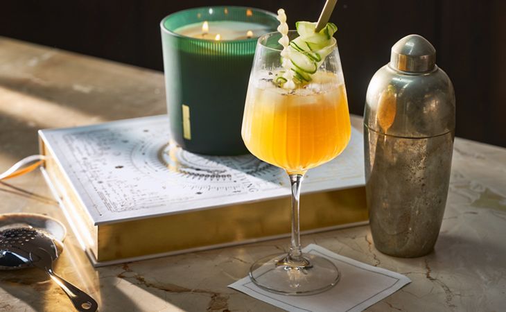 Cóctel sin alcohol inspirado en The Ritual of Jing, con pepino de decoración, al lado de una vela aromática a juego y una coctelera vintage.