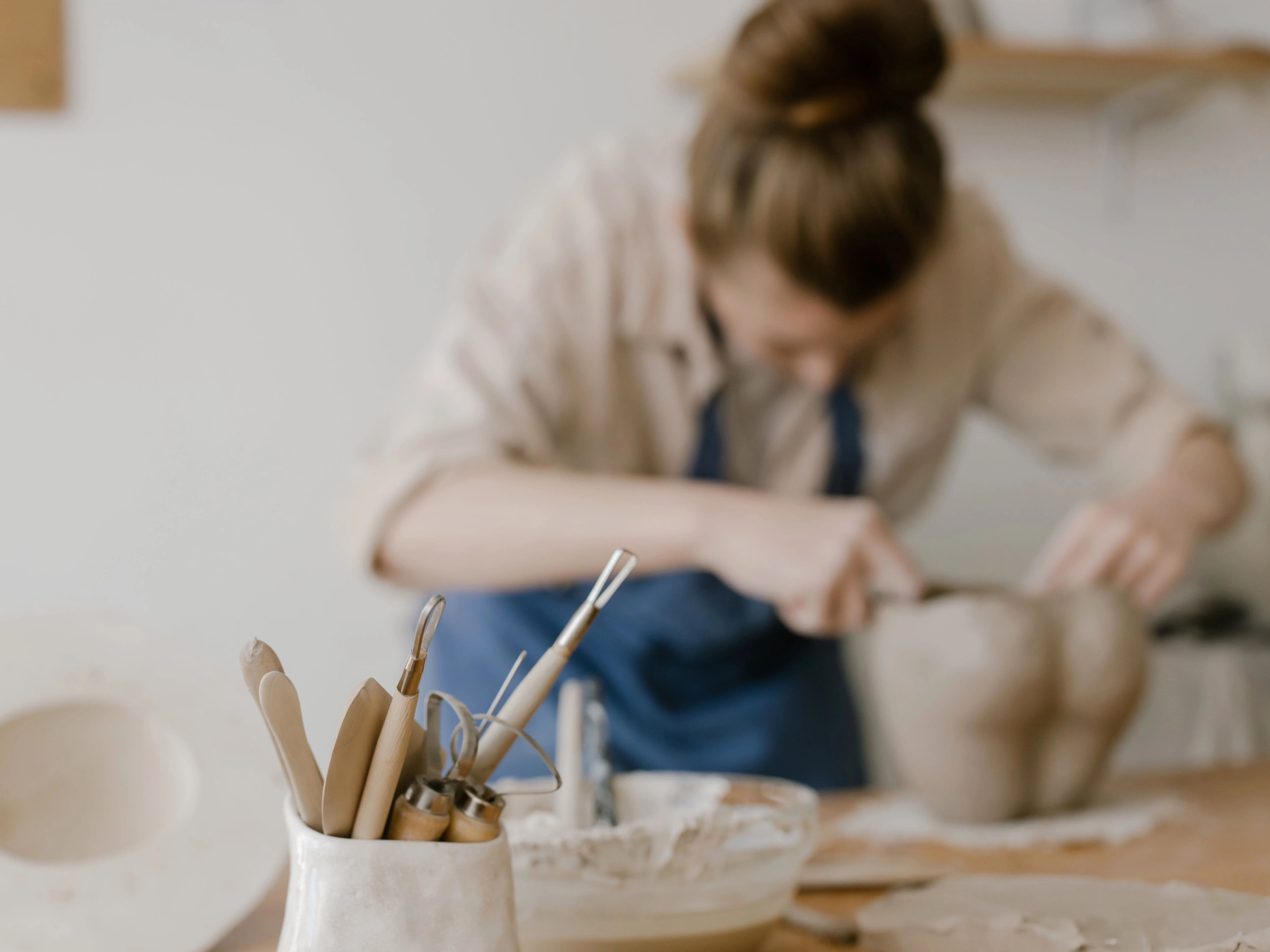 Woman shaping clay while pottery tools in focus on a worktable in the foreground.